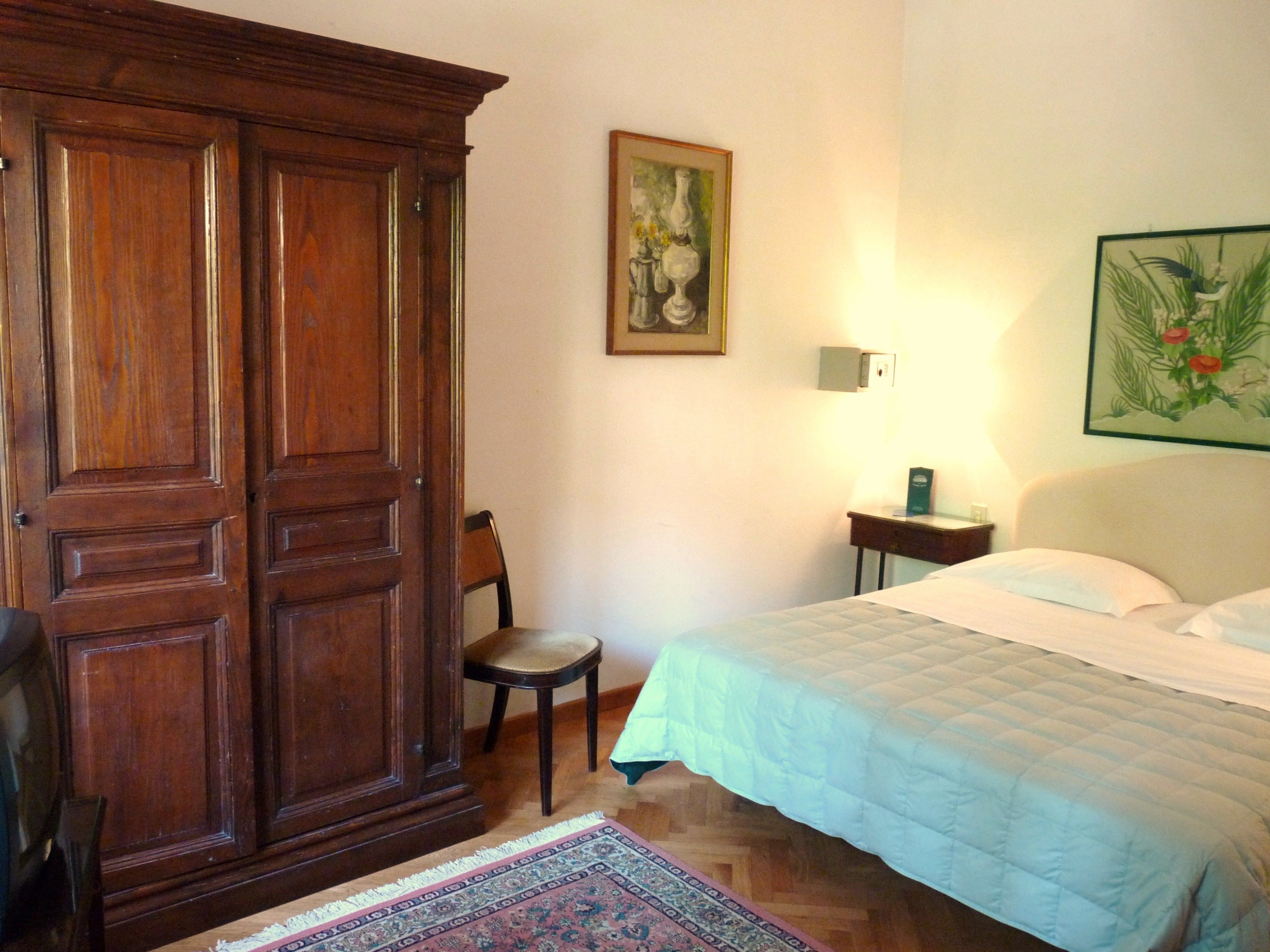A bedroom with a bed, a wooden wardrobe, a chair, a nightstand, and a wall-mounted lamp.
