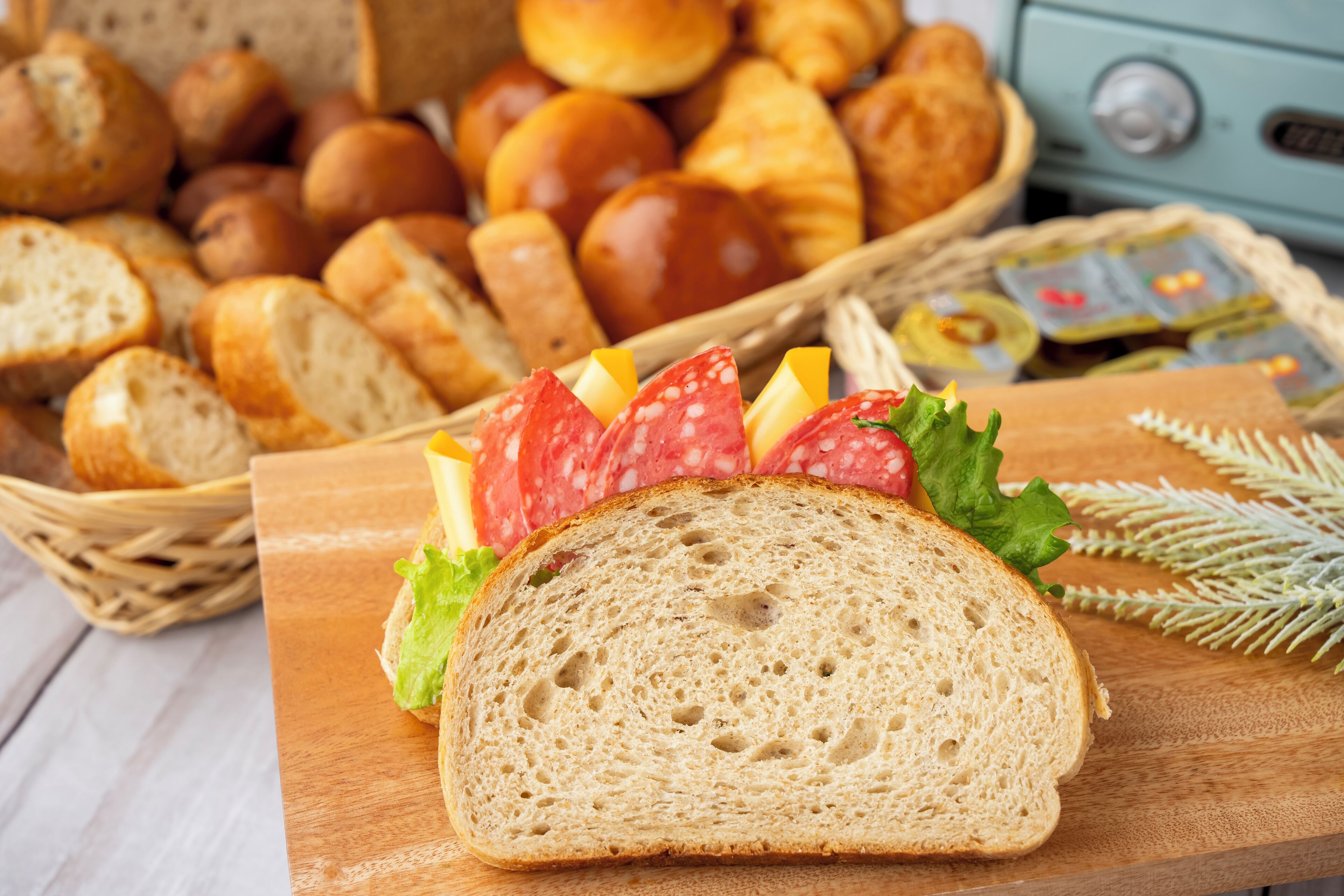 Una rebanada de pan con queso y salame sobre una tabla de madera, rodeada de diferentes tipos de pan y pasteles.