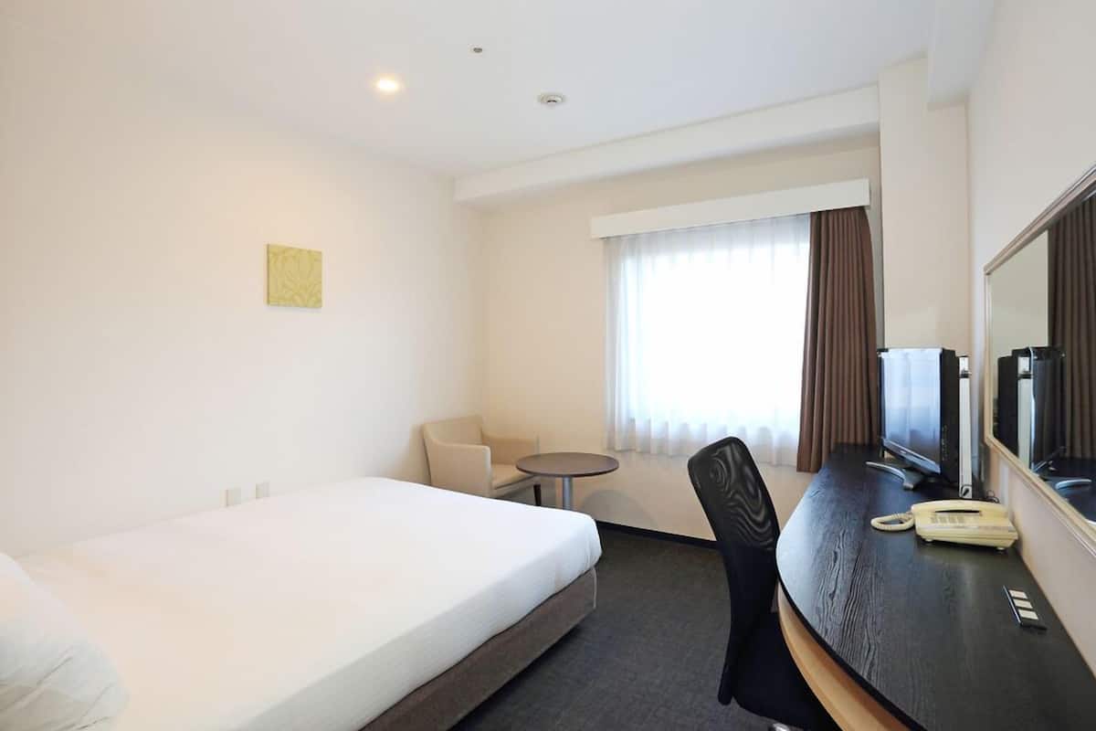 A hotel room with a bed, a desk with a telephone, a chair, a small table, and a window with curtains.