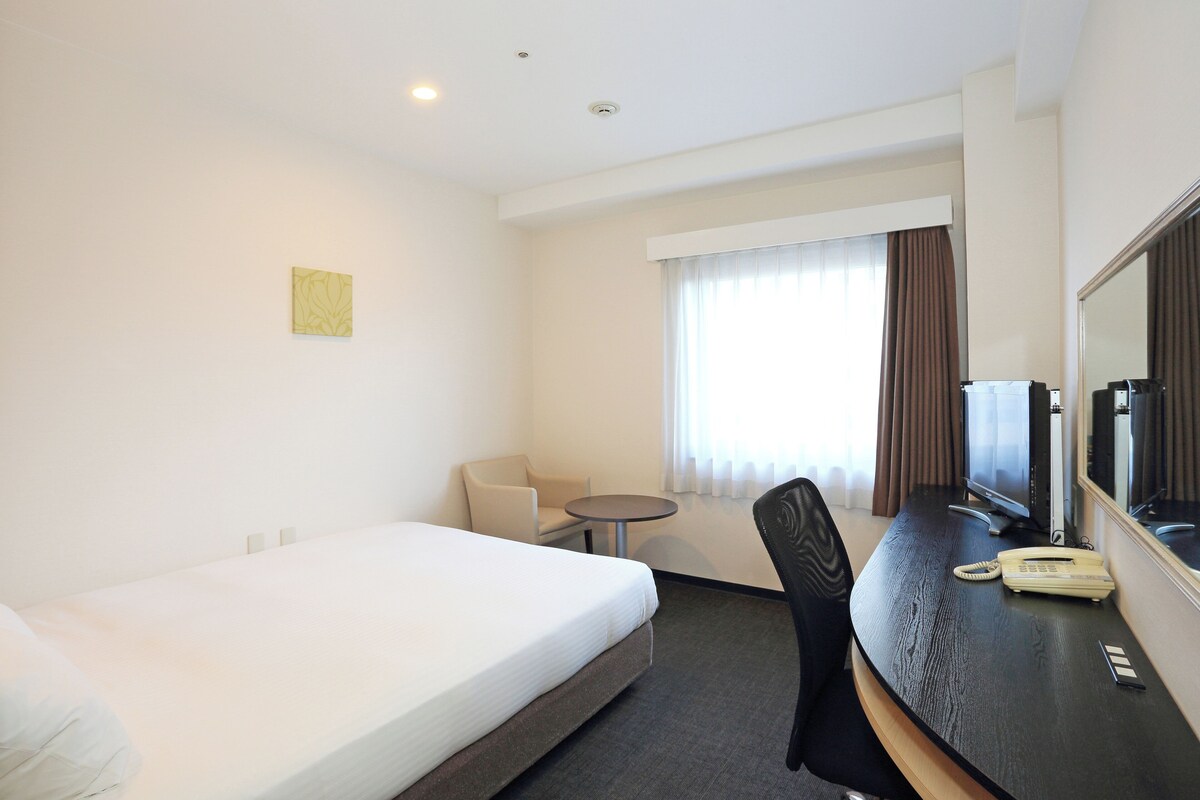 A hotel room with a single bed, a desk with a telephone, a chair, a small table, and a window with curtains.