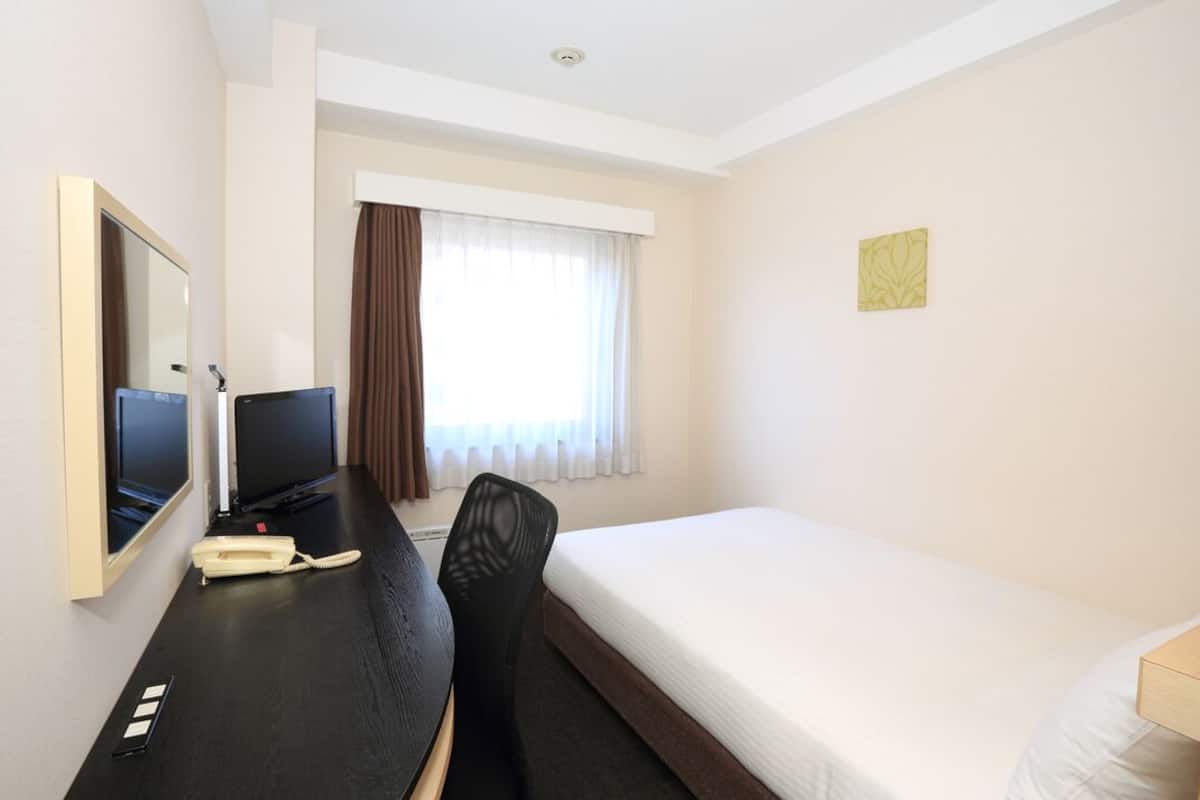 A hotel room with a bed, a desk with a telephone, a chair, a television, and a window with curtains.