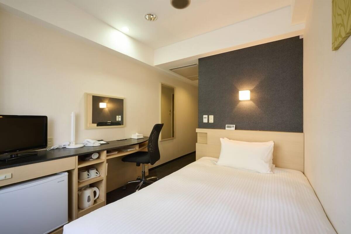 A hotel room with a bed, a desk with a chair, a television, a mini-fridge, and a wall-mounted light.