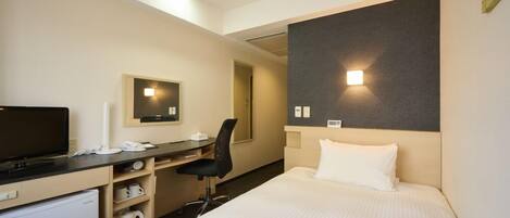 A hotel room with a bed, a desk with a chair, a television, a mini-fridge, and a wall-mounted light.