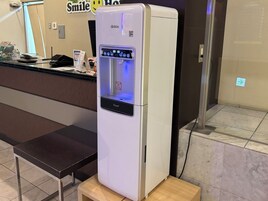 Um dispensador de água branca com visor digital, posicionado sobre uma plataforma de madeira em ambiente comercial.