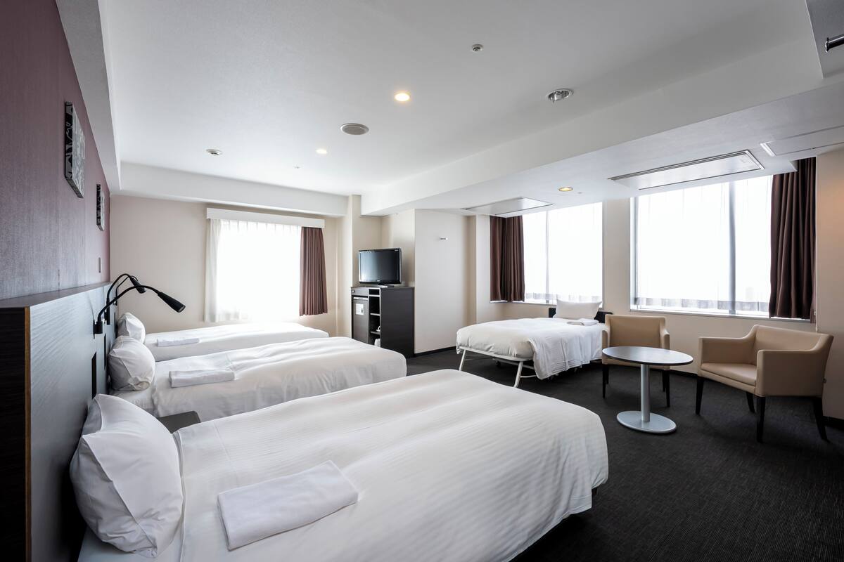 A hotel room with two beds, a desk, a chair, a TV, and large windows.