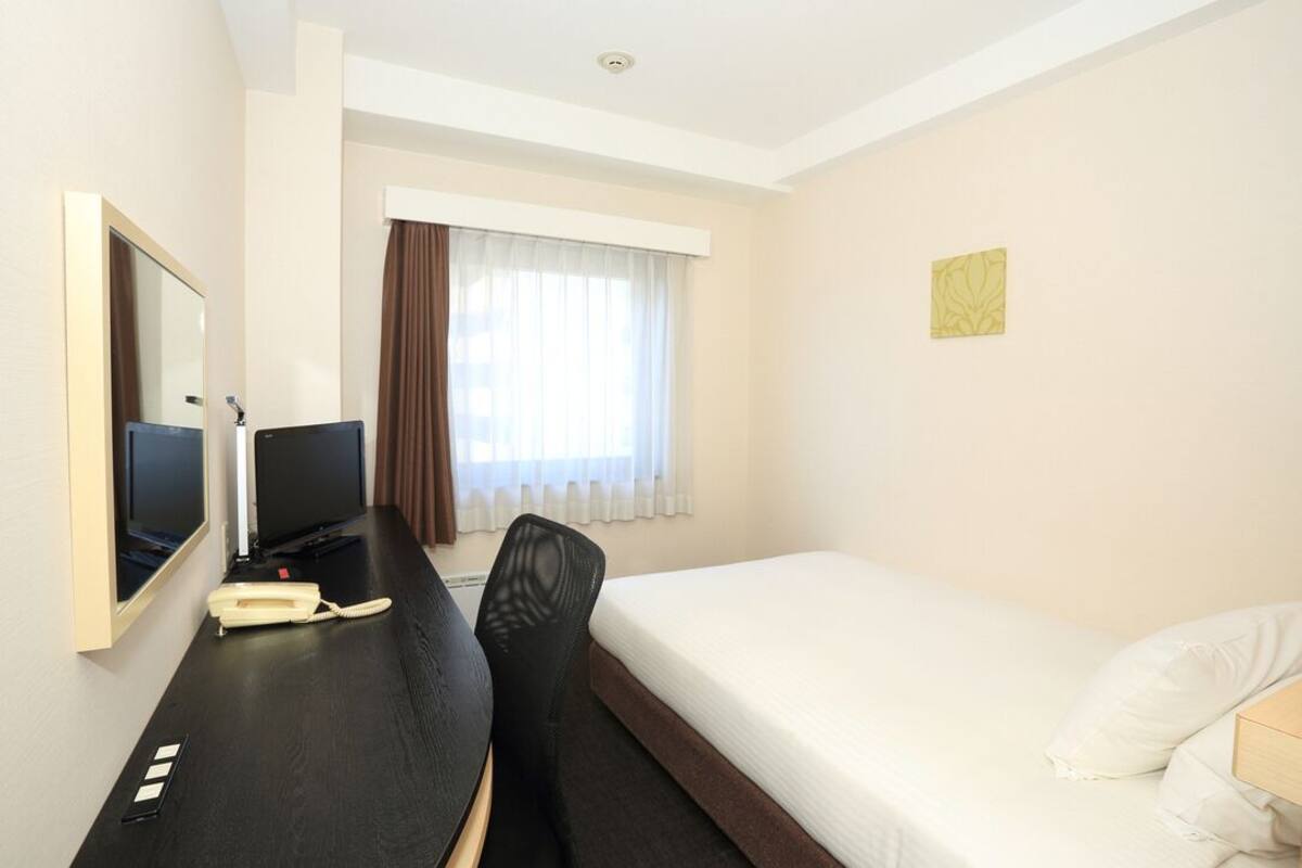 A hotel room with a bed, a desk with a telephone, a chair, a television, and a window with curtains.