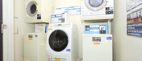 Un cuarto de lavado con varias lavadoras y secadoras, incluyendo una lavadora de carga frontal y otra de carga superior. Se observan instrucciones y etiquetas en los equipos y paredes.