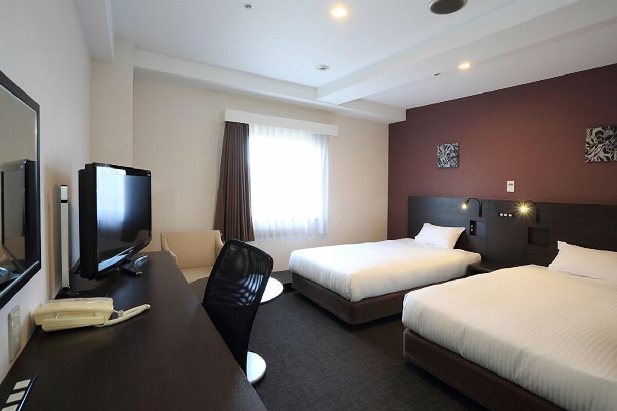 A hotel room with a bed, a desk with a television, a chair, and a window with curtains.