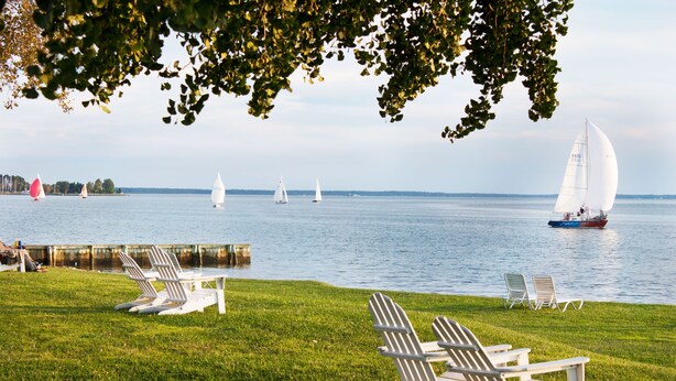 Eine Rasenfläche mit weißen Adirondack-Stühlen und Blick auf Segelboote auf dem Wasser.