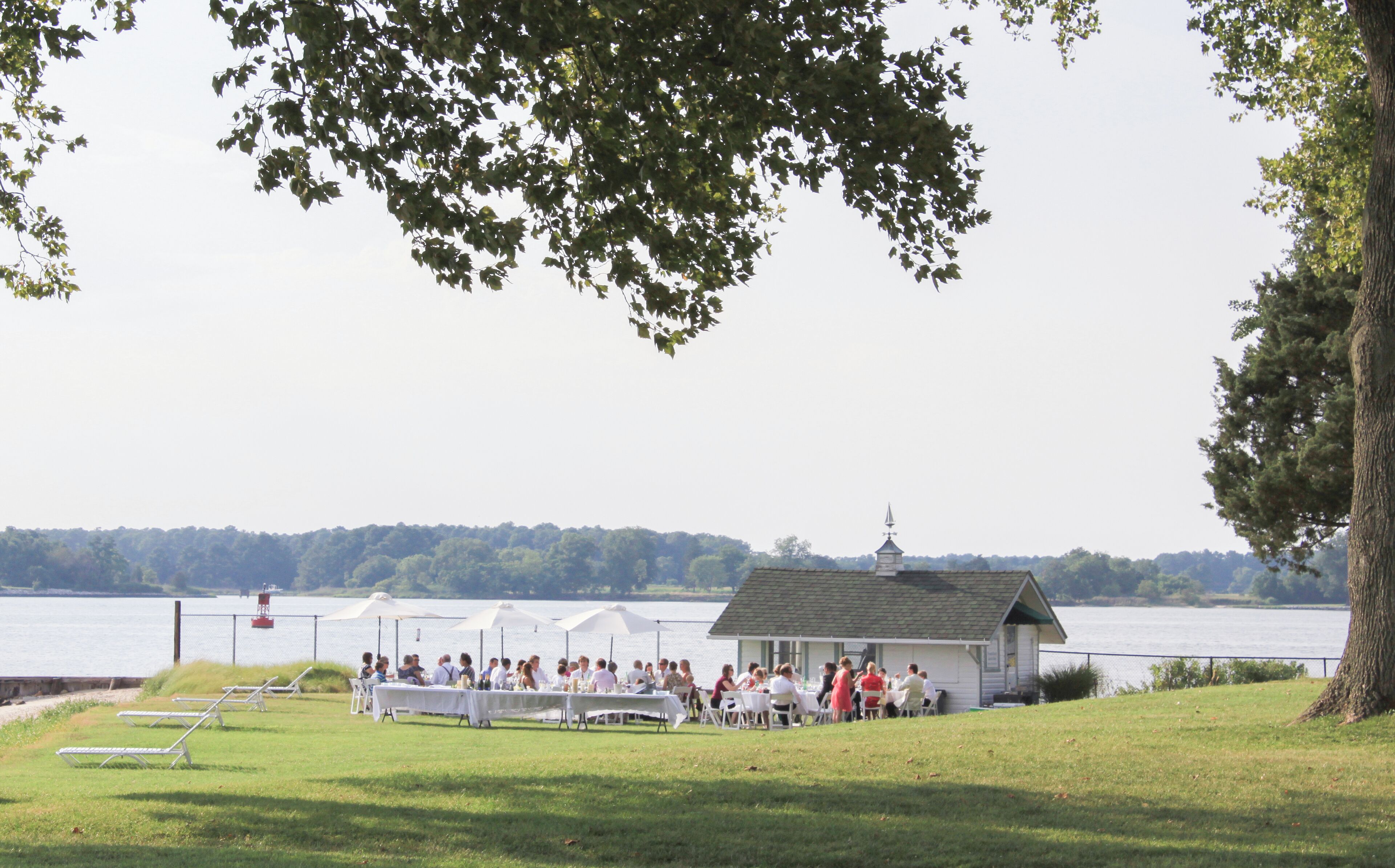 Evento junto al lago con mesas y sillas dispuestas para los invitados, un pequeño edificio y vistas al lago y a los árboles.