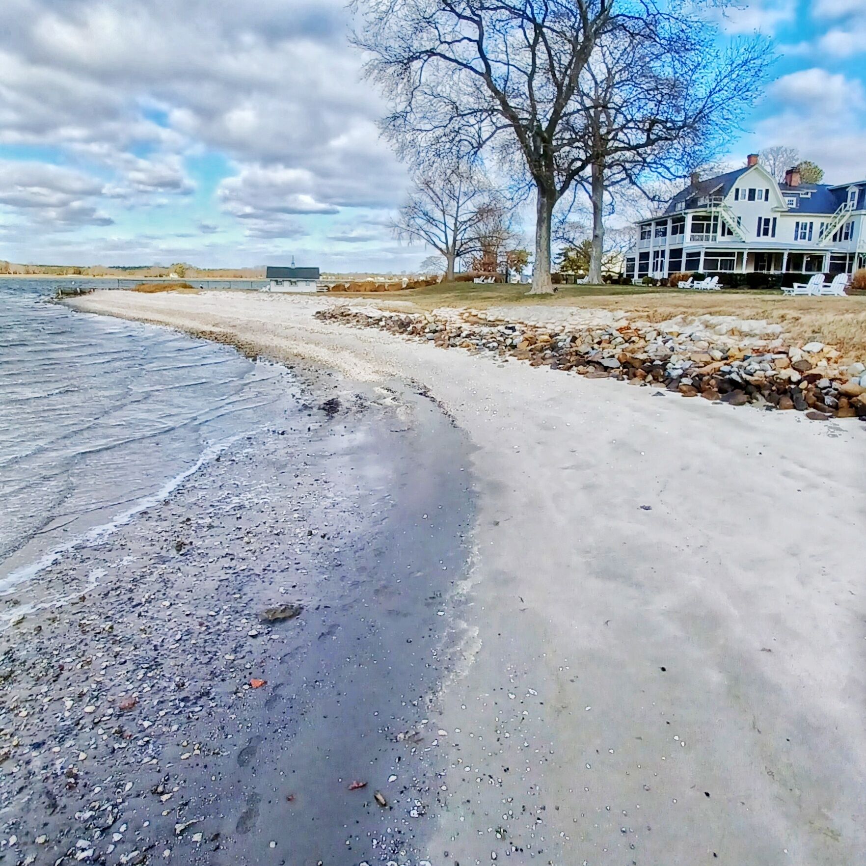 A waterfront property with a sandy beach, a rocky shoreline, and a large house with a porch.