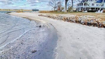 A waterfront property with a sandy beach, a rocky shoreline, and a large house with a porch.