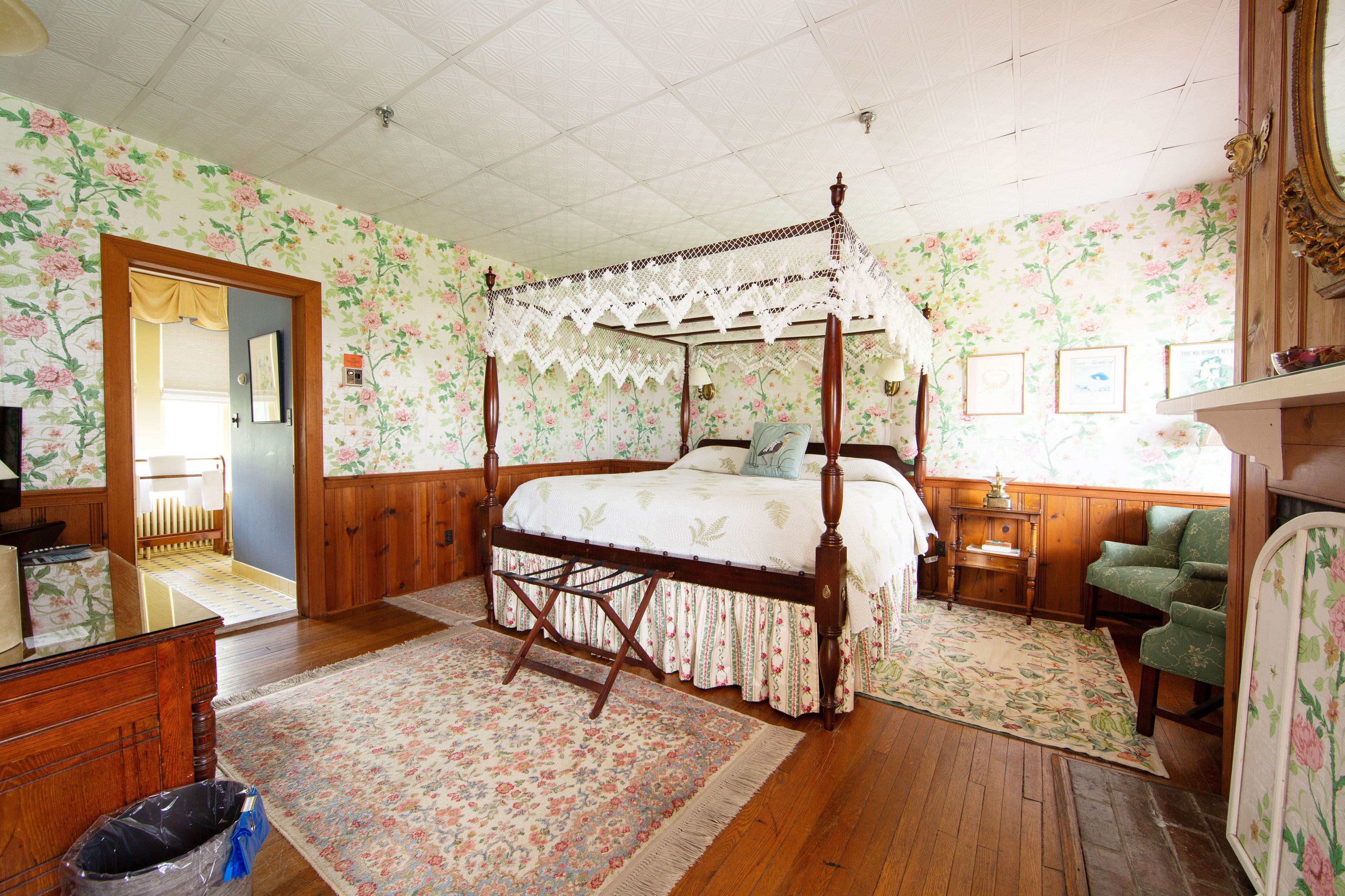 A four-poster bed with floral wallpaper, a wooden floor, a green armchair, and a mirror.
