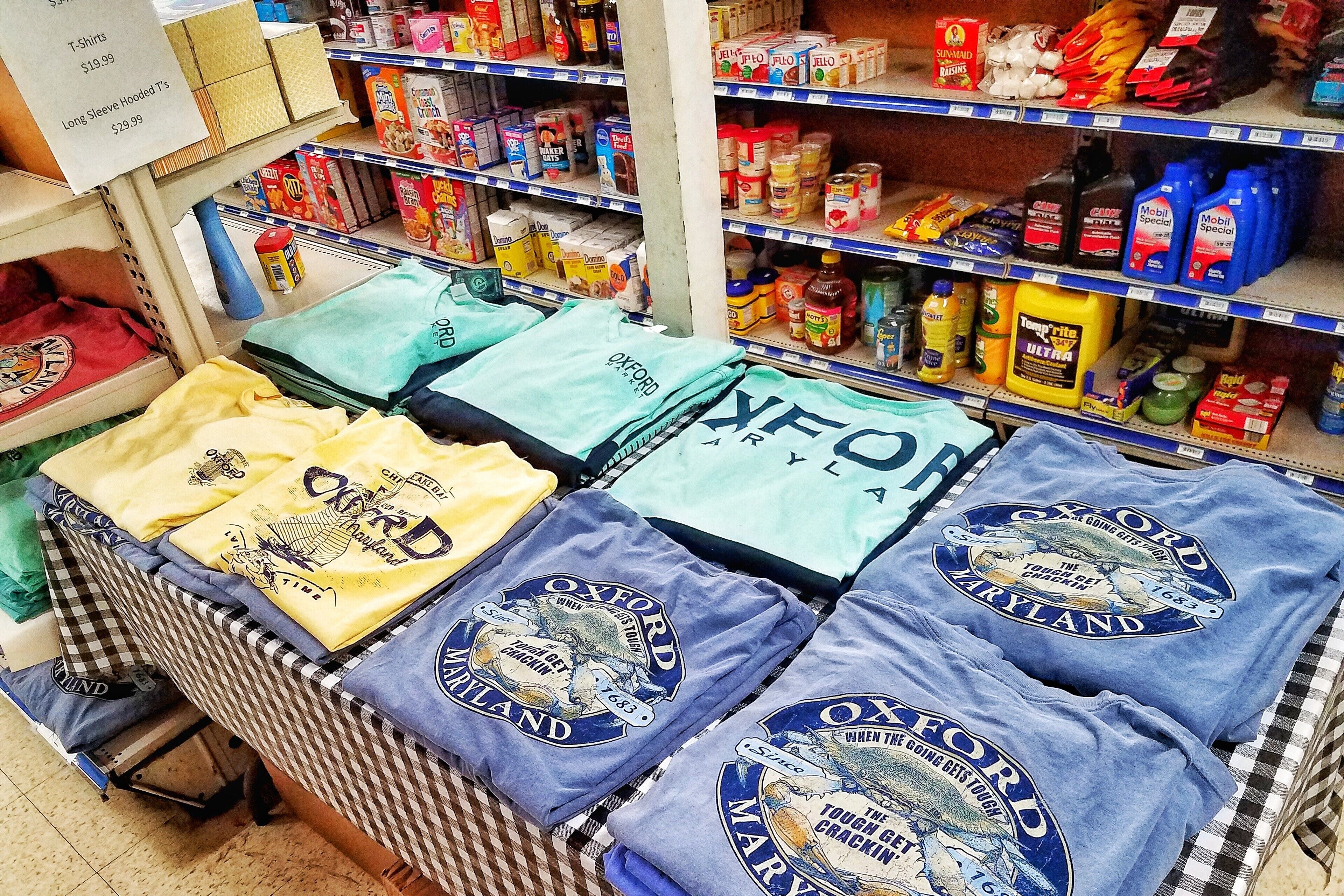 Una tienda minorista con estantes repletos de diversos productos, incluyendo camisetas, aperitivos y artículos de limpieza.