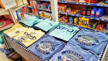 Una tienda minorista con estantes repletos de diversos productos, incluyendo camisetas, aperitivos y artículos de limpieza.
