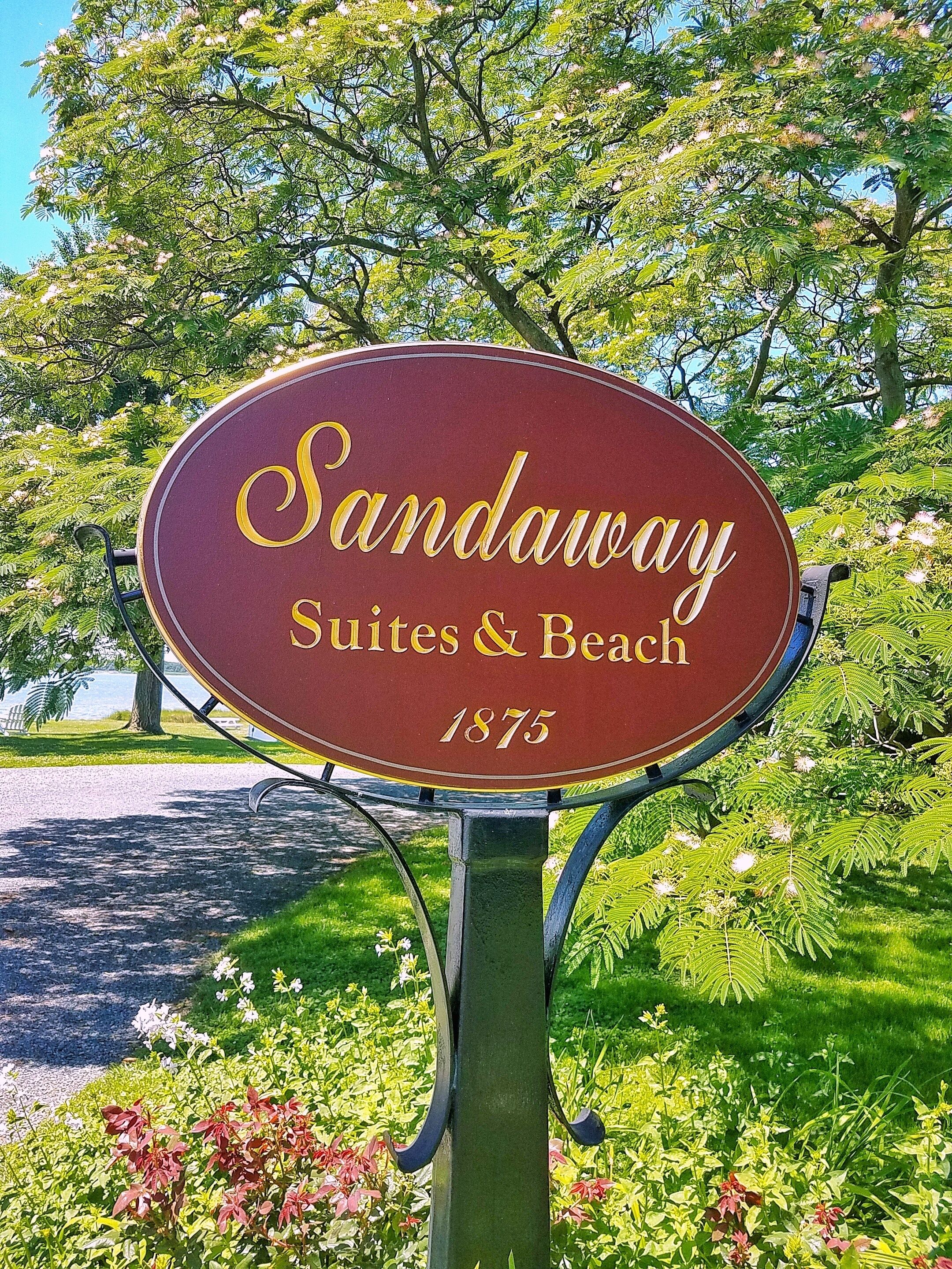 A sign for Sandaway Suites & Beach, established in 1875, with a clear view of the surrounding greenery and a pathway.