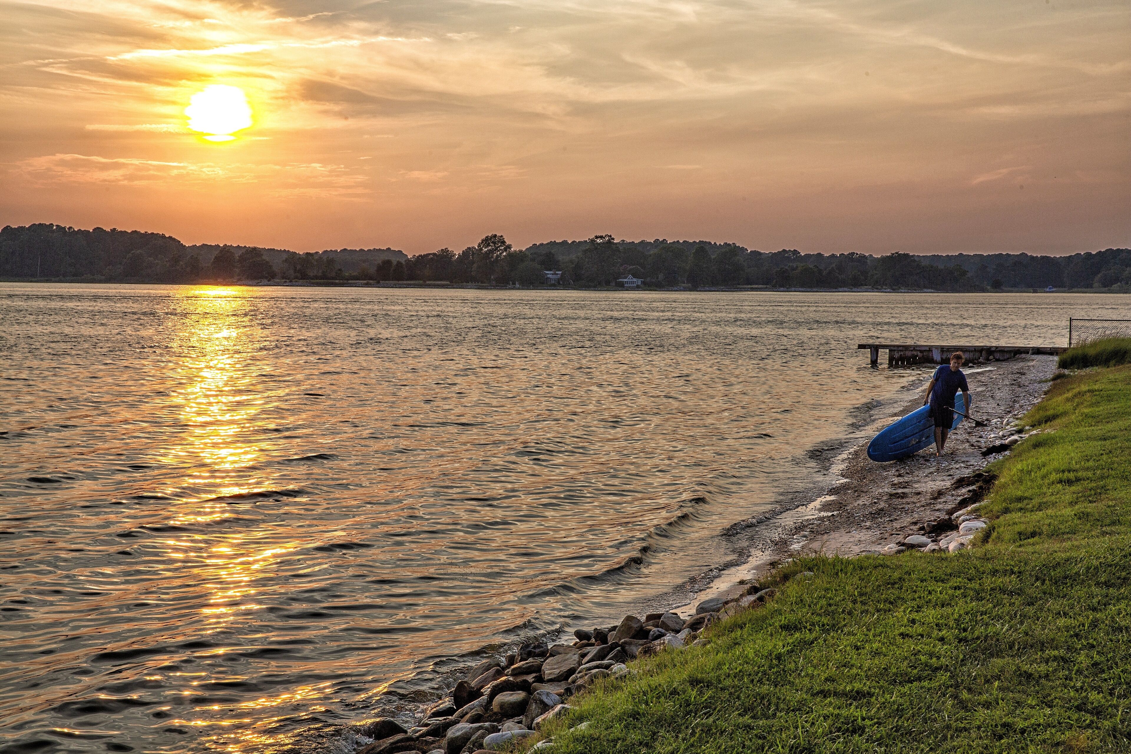 Tramonto su un lago, con una persona che trasporta un'asse da surf vicino a un pontile.