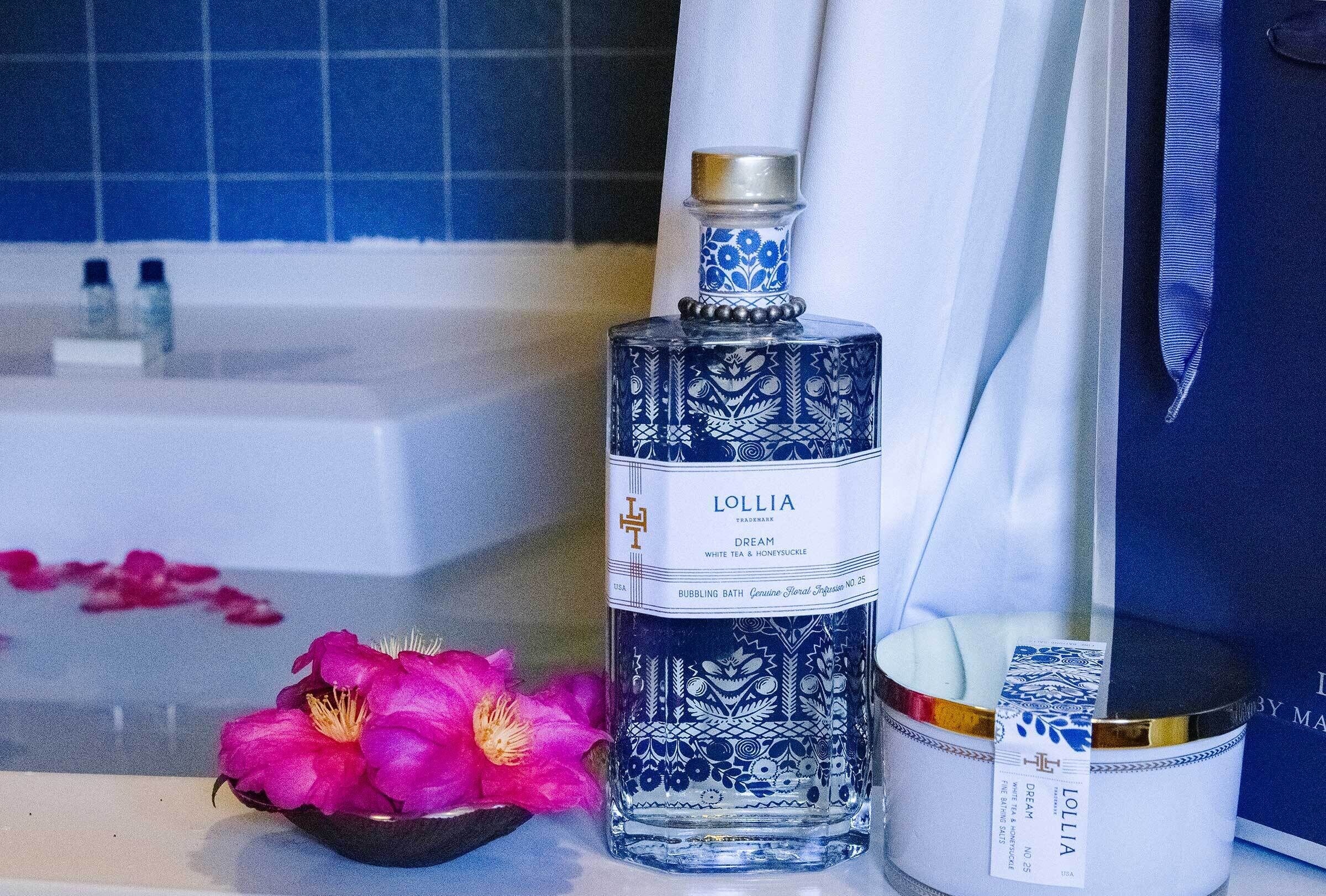 A bottle of Lollia Dream white tea and honey syrup on a bathroom counter with a bathtub and pink flowers.