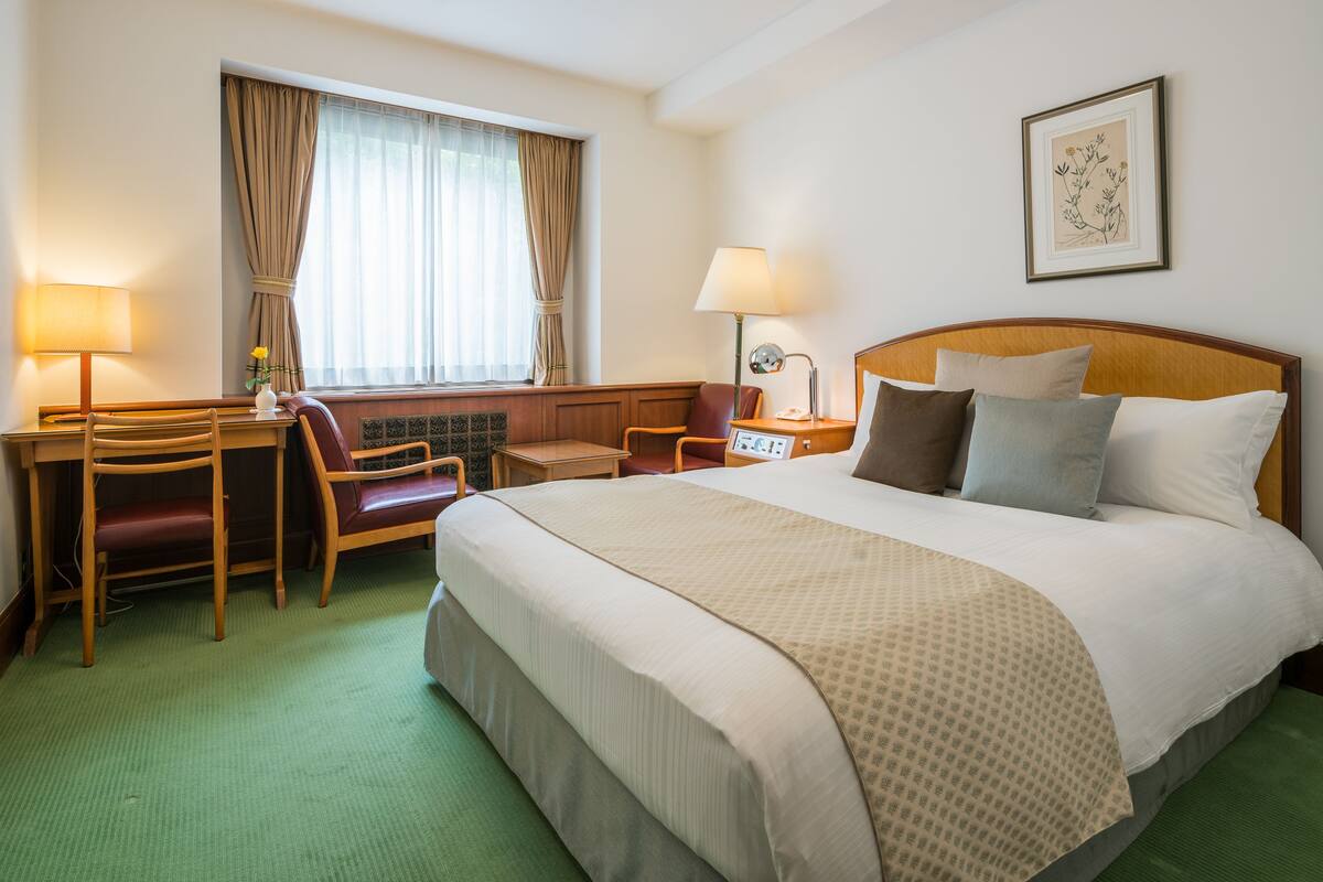 A hotel room with a bed, a desk, a chair, a lamp, and a window with curtains.