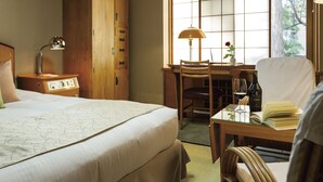 A hotel room with a bed, a desk, a chair, a lamp, a clock, and a window with a view of trees. - Hilltop Hotel (Tokyo)