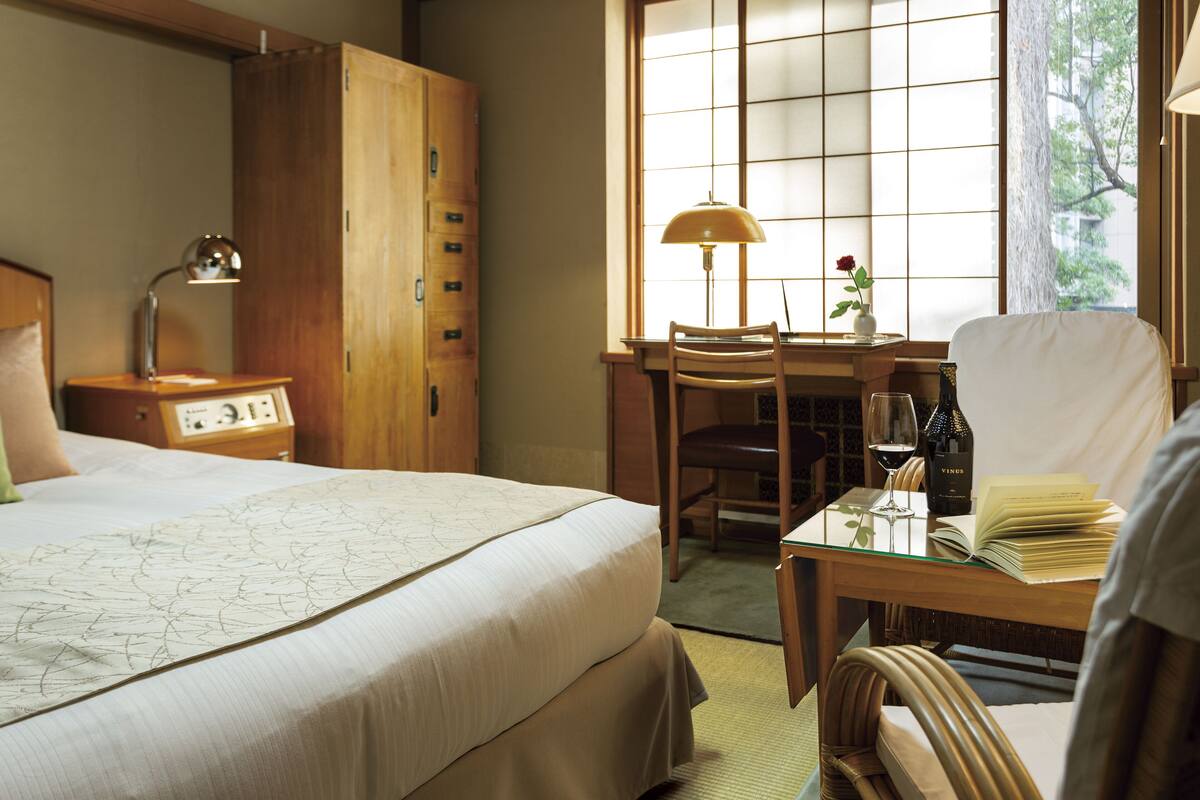 A hotel room with a bed, a desk, a chair, a lamp, a clock, and a window with a view of trees.