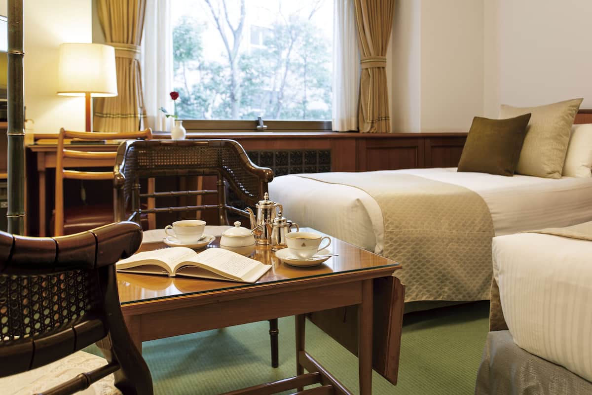 A hotel room with a bed, a table with a book and tea set, a chair, and a window with curtains.