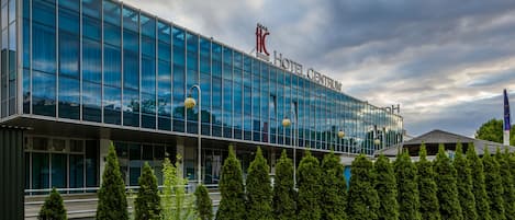 A modern hotel building with a glass facade, a prominent logo, and a well-maintained garden in front.