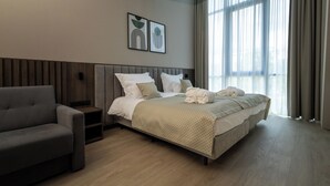 Une chambre d’hôtel moderne avec un grand lit, un canapé et une table de chevet. Deux tableaux encadrés ornent le mur, et la chambre dispose d’une grande fenêtre avec des rideaux.