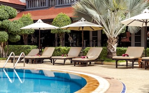 A pool area with lounge chairs and umbrellas, surrounded by greenery and a building with a red-tiled roof. - Lucky Angkor Hotel & Spa (Siem Reap)
