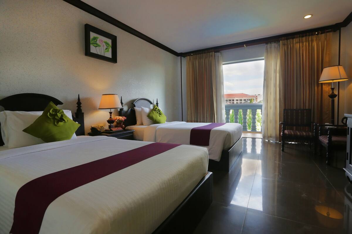 A hotel room with two beds, a nightstand with a lamp, a chair, and a view of the outside.