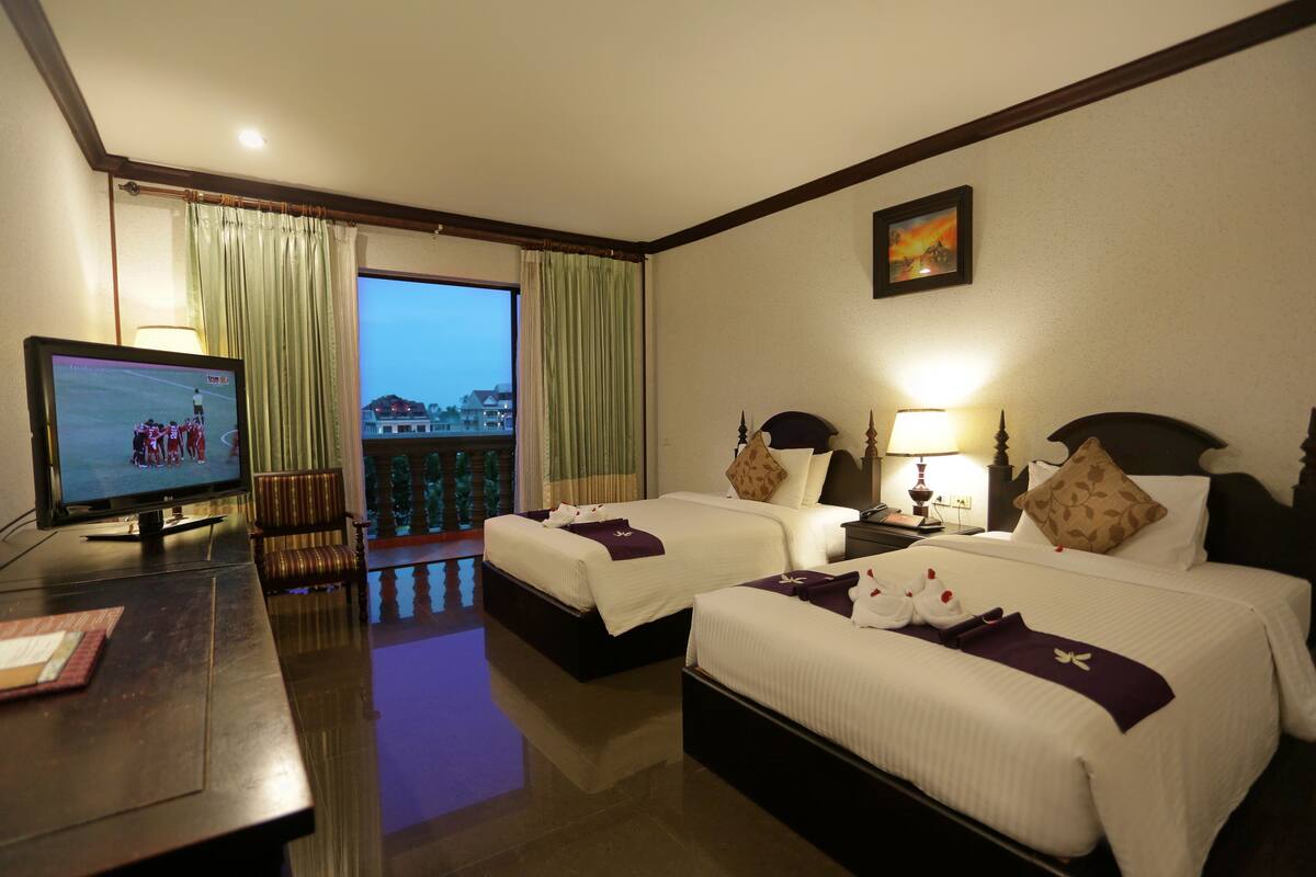 A hotel room with two beds, a TV, a desk, and a view of a building through the window.