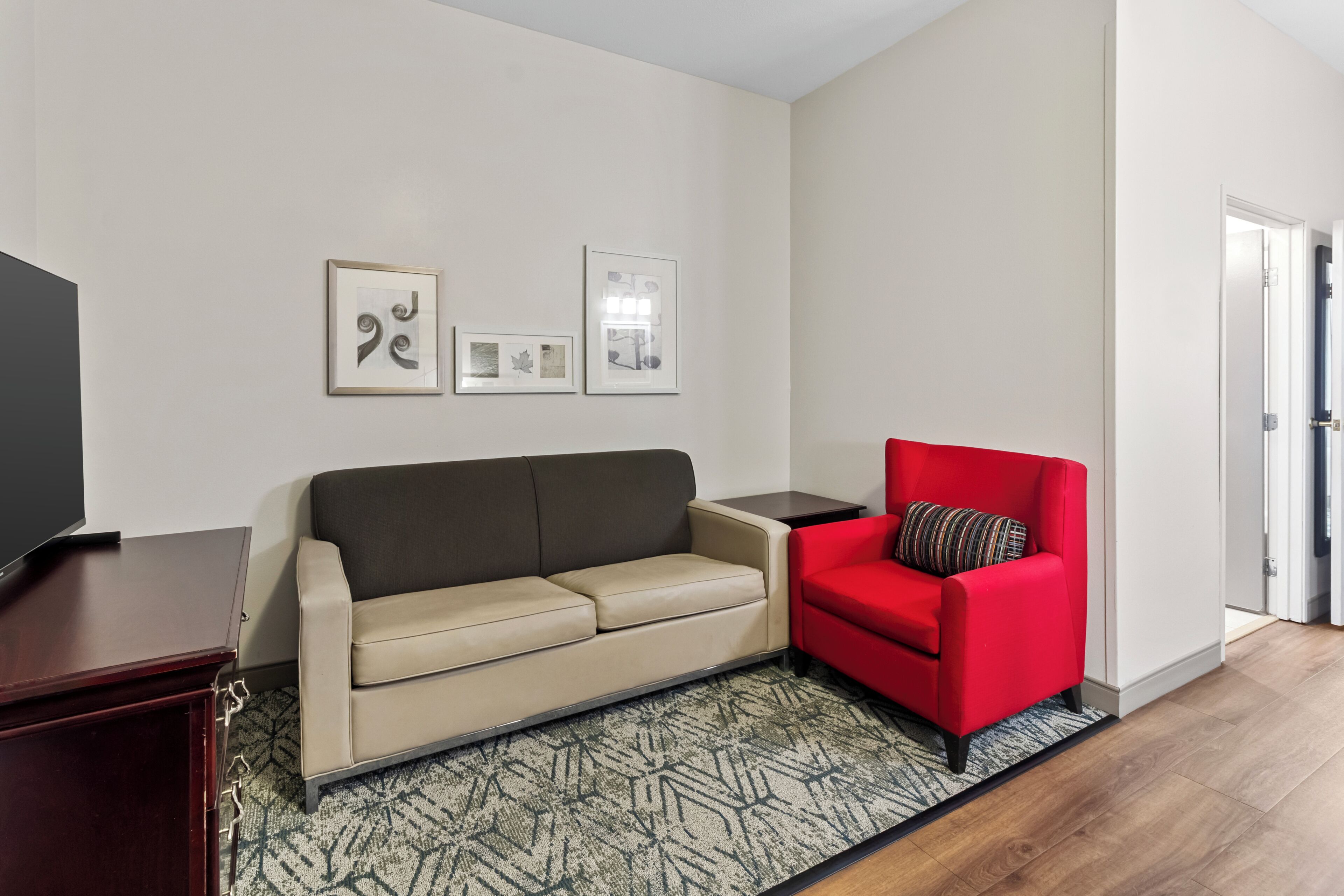A living room with a beige sofa, a red armchair, a TV on a stand, and framed pictures on the wall.