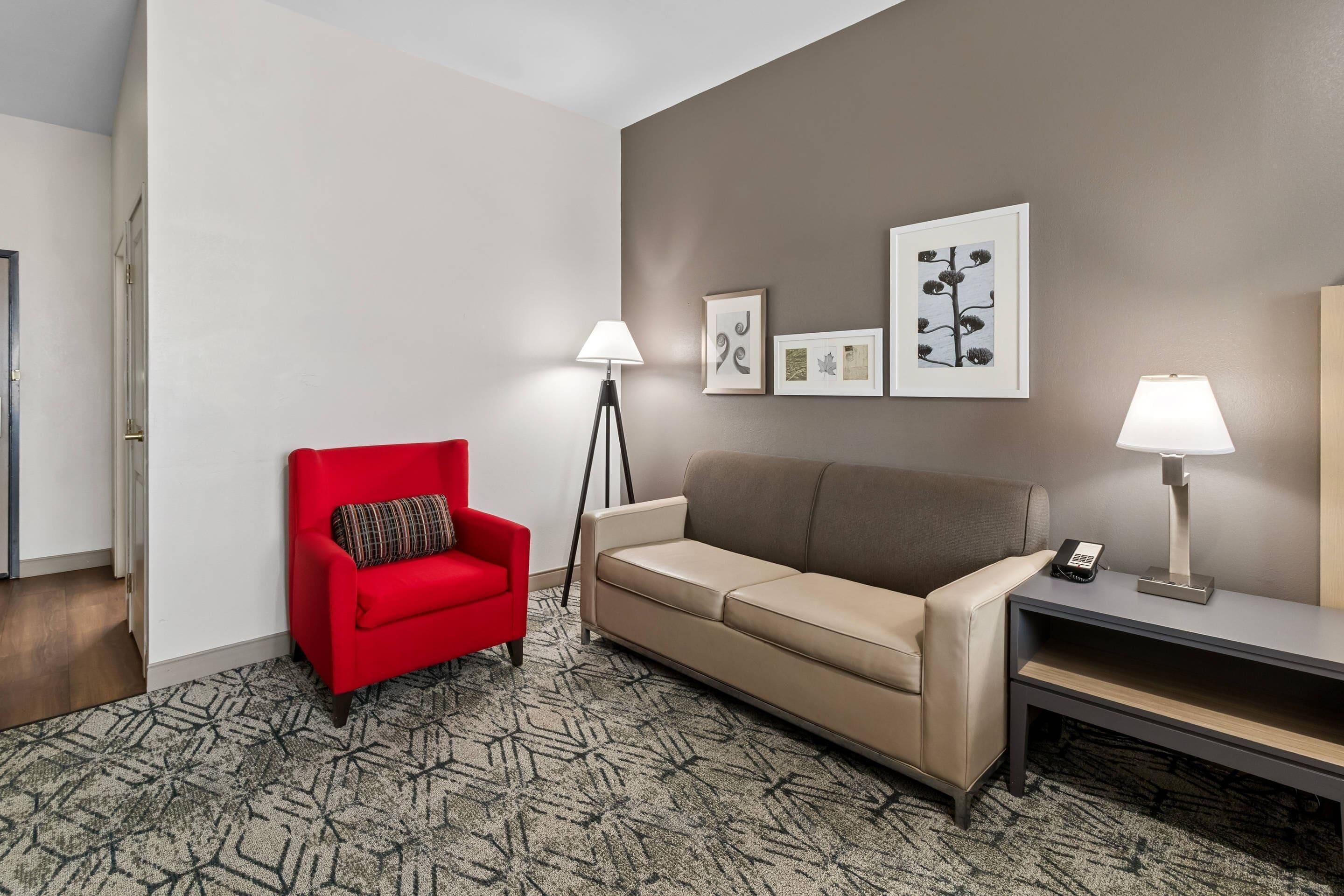 Habitación de hotel con un sillón rojo, un sofá color beige, una lámpara de pie, una mesita de noche con un teléfono y obras de arte enmarcadas en la pared.