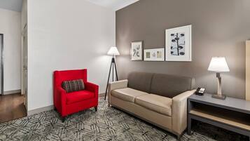 Habitación de hotel con un sillón rojo, un sofá color beige, una lámpara de pie, una mesita de noche con un teléfono y obras de arte enmarcadas en la pared.