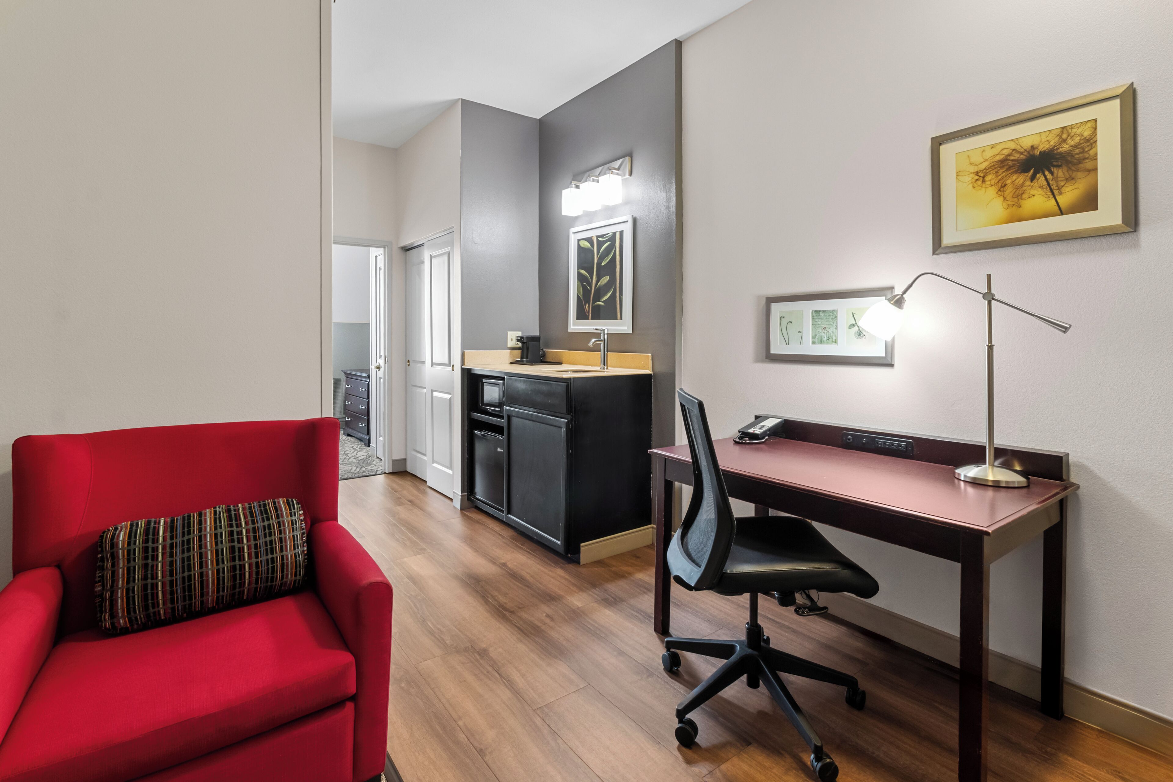 Et moderne hotelværelse med en rød lænestol, et skrivebord med lampe, et tekøkken og et spisebord.