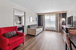 Habitación de hotel con un sofá rojo, un sofá color crema, una mesa de comedor con sillas, un televisor y un ventanal grande con cortinas.
