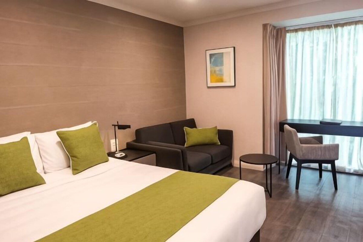 Una habitación de hotel con cama, sofá, sillón, escritorio y ventana con cortinas.