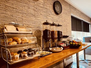 Een ontbijtbuffet met diverse gebakjes en fruit, een klok aan de muur, en een raam met zonwering.
