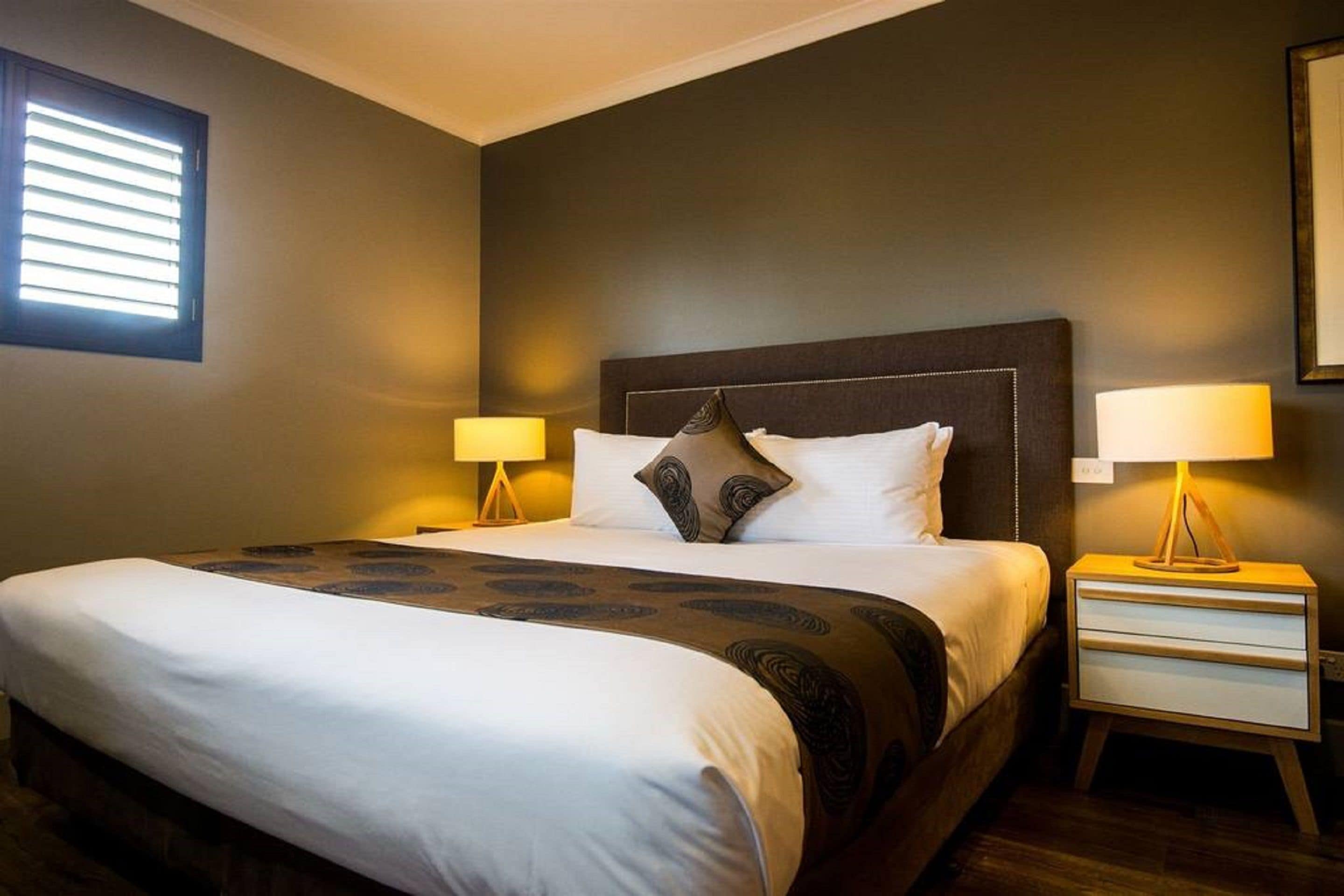 Habitación de hotel con una cama grande, dos mesitas de noche con lámparas, una ventana con persianas y una pared de color oscuro.