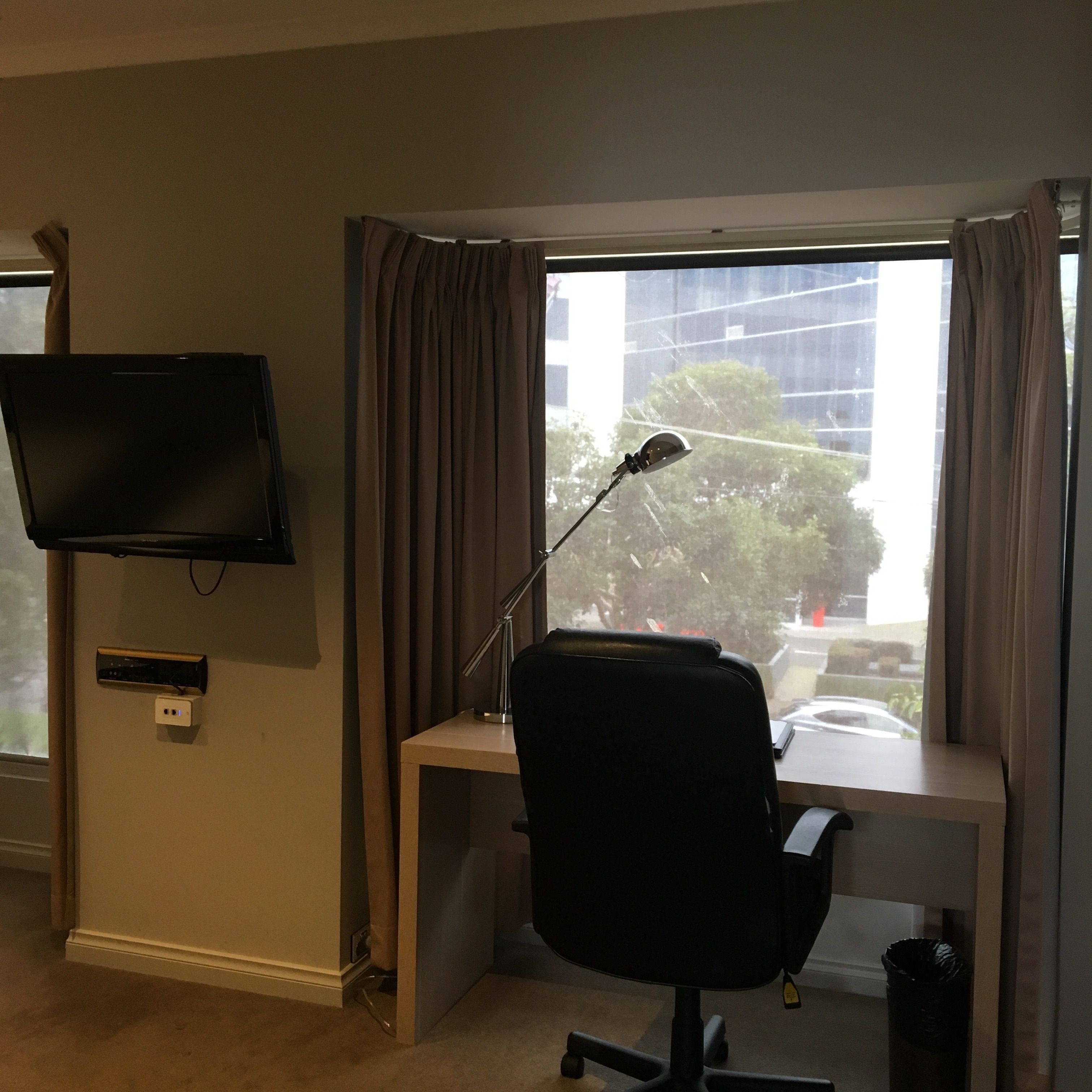 A hotel room with a desk, a chair, a TV mounted on the wall, and a window with curtains.