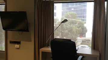 Ein Hotelzimmer mit Schreibtisch, einem Sessel, einem an der Wand befestigten Fernseher und einem Fenster mit Vorhängen.