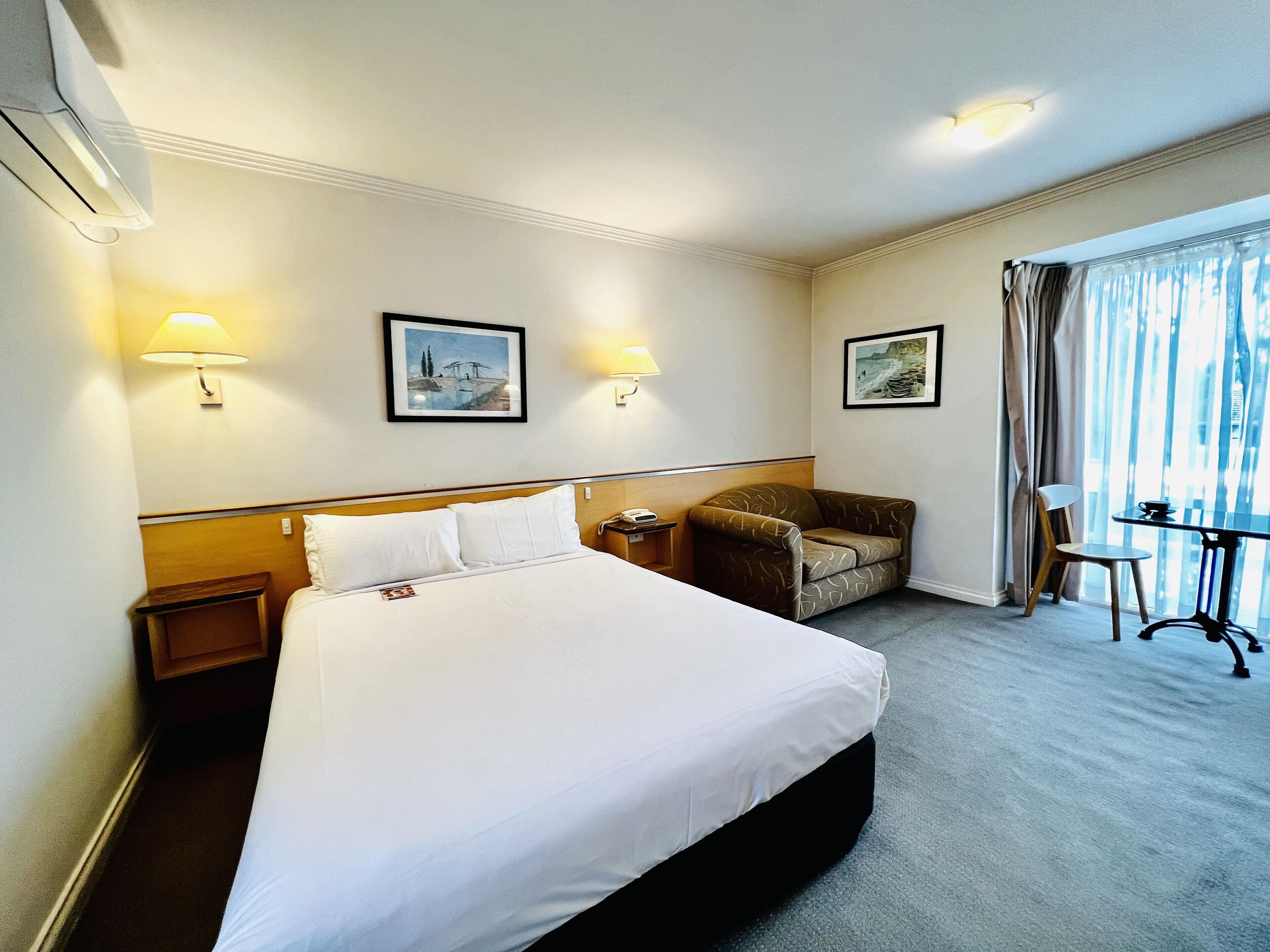 A hotel room with a large bed, two bedside lamps, a chair, a small table, and a window with curtains.