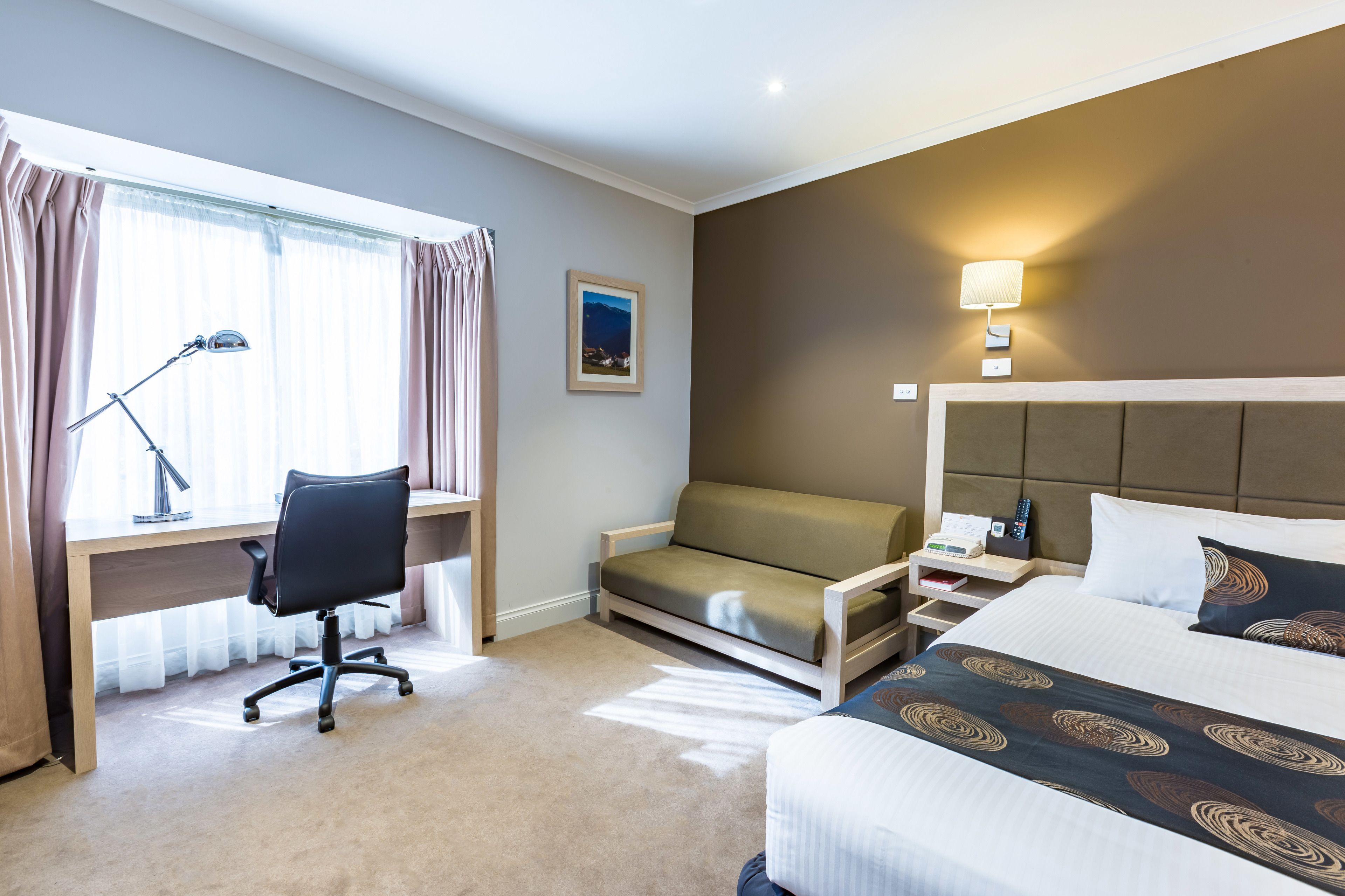 Habitación de hotel con cama, escritorio con silla, sofá, mesita de noche con lámpara y televisor montado en la pared.