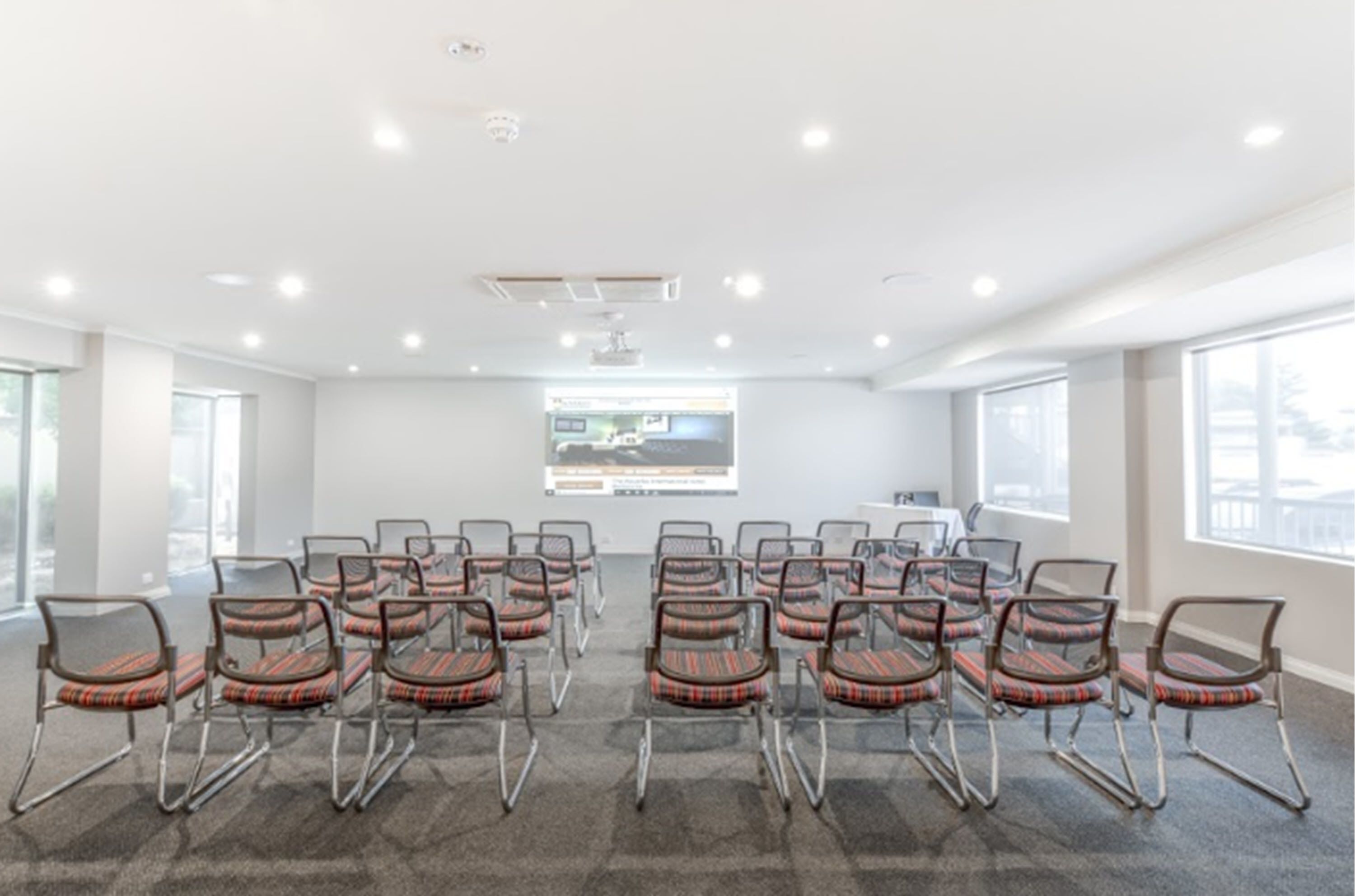 A conference room with rows of chairs facing a presentation screen, carpeted floor, and large windows.