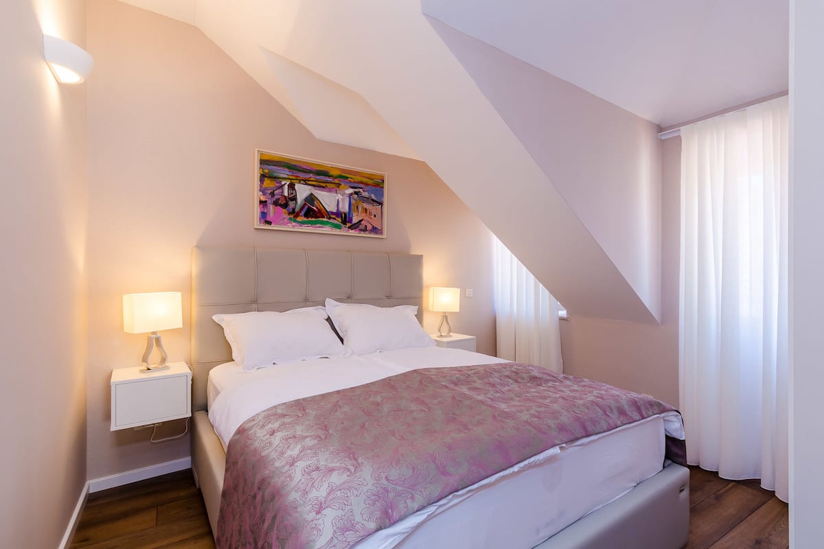 Ein Schlafzimmer mit einem Bett, zwei Nachttischen, einem Gemälde und einem Fenster mit Vorhängen.