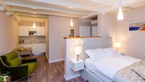 A compact hotel room with a bed, bedside table, green chair, kitchenette, and a painting on the wall. - Villa Flores - Apartments (Dubrovnik)