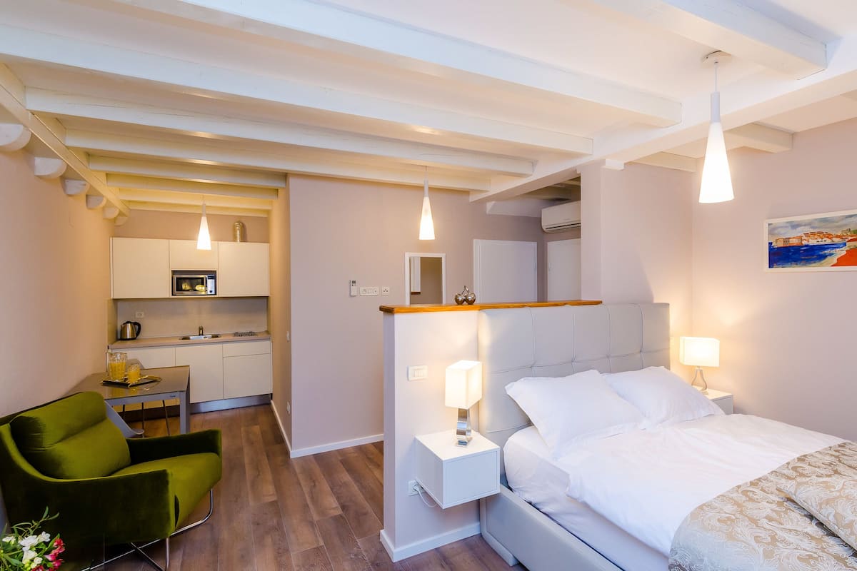 Ein kleines Hotelzimmer mit Bett, Nachttisch, grünem Sessel, einer Küchenzeile und einem Bild an der Wand.