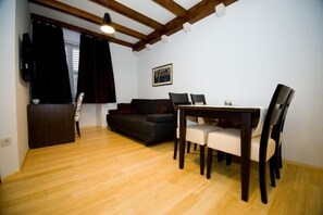 A living room with a wooden floor, a dark sofa, a dining table with chairs, and a TV mounted on the wall.