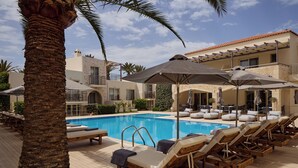 Zona de piscina con sillas de descanso y sombrillas, rodeada de palmeras y un edificio con balcones.