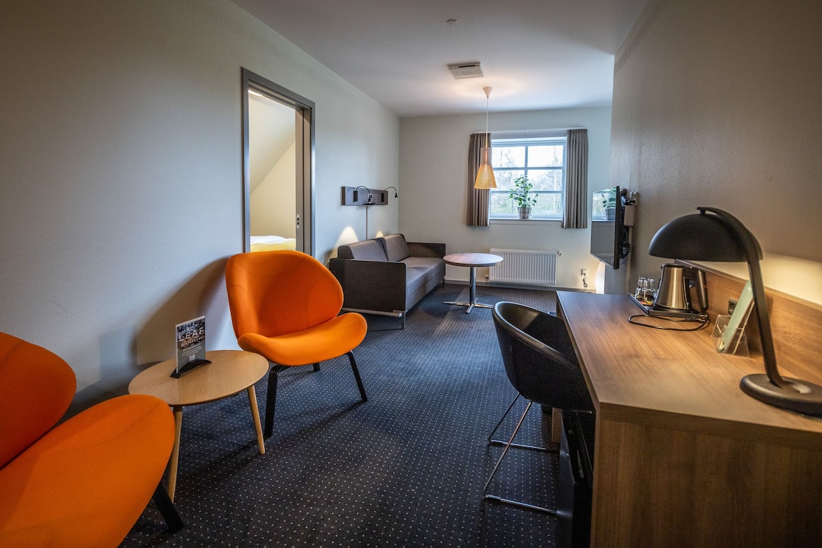 Moderni hotellihuone, jossa on sohva, kaksi oranssia nojatuolia, pieni pyöreä pöytä, työpöytä lampun kanssa ja ikkuna, jossa on verhot.